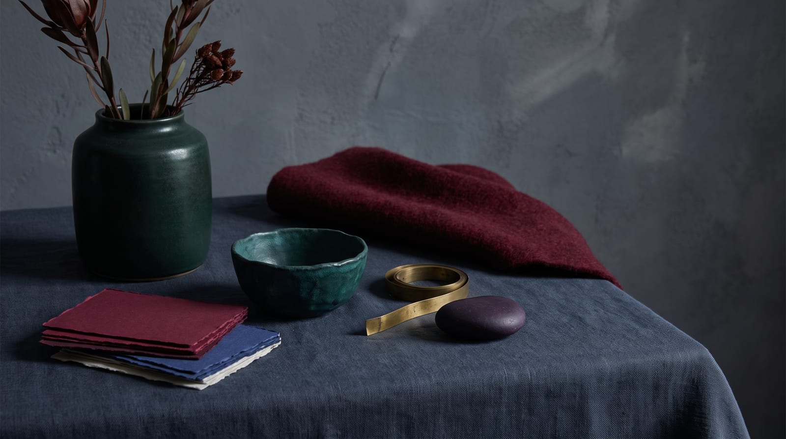 A deep winter editorial still-life: a high-contrast ensemble of folded jet-black wool, a faceted emerald-green glass vessel, a stem of dark-berried holly, and a shard of icy quartz on black slate, every element rendered in the palette's near-blacks, cold jewel tones, and one bright icy accent.