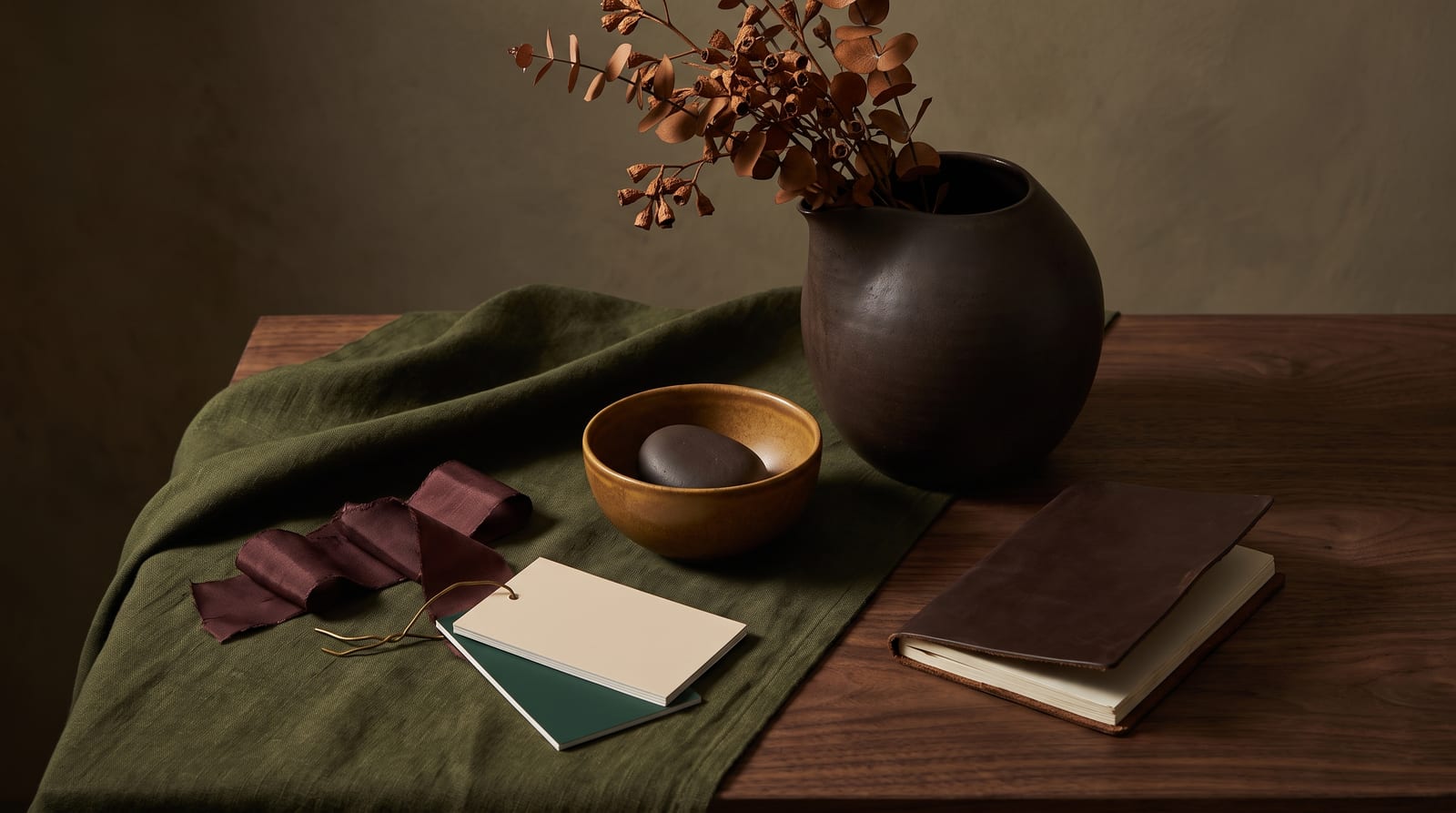 A deep autumn editorial still-life: a tactile ensemble of folded chocolate-brown wool, a burnished bronze vessel, a cluster of dried rust-red seedheads, and a piece of dark walnut on slate, every element rendered in the palette's espressos, rusts, deep olives, and burnished golds.