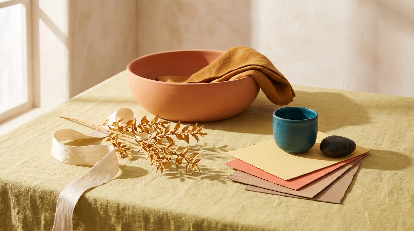 A warm spring editorial still-life: a tactile ensemble of folded golden-yellow linen, a small glazed coral ceramic vessel, a stem of fresh green leaves, and an apricot-toned stone on warm cream plaster, every element rendered in the palette's golden yellows, corals, warm turquoises, and leaf greens.