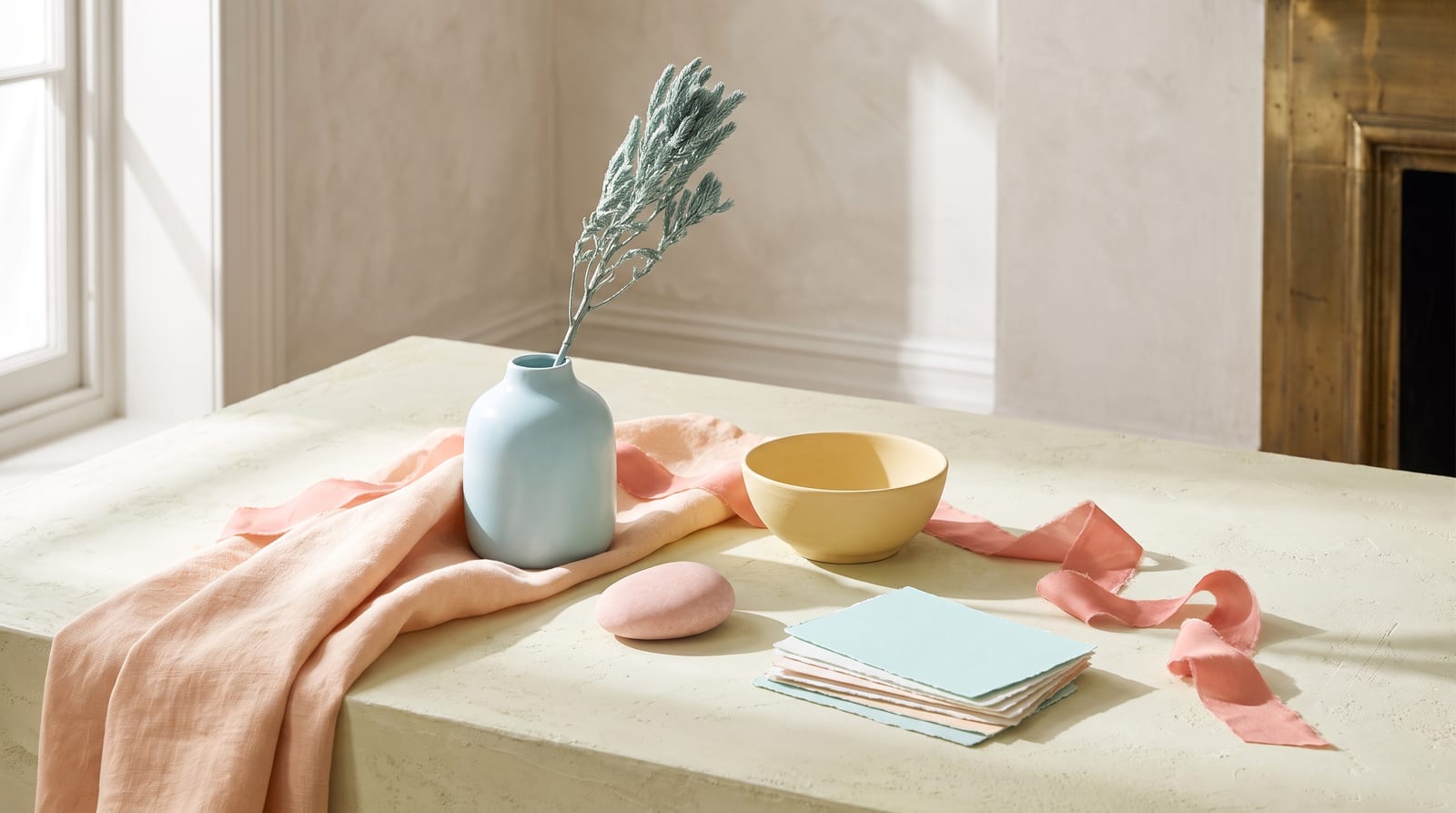 A light spring editorial still-life: a tactile ensemble of folded peach linen, a small glazed apricot-toned ceramic cup, a stem of fresh freesia, and a smooth warm-cream pebble on pale sunlit plaster, every element rendered in the palette's peaches, corals, golden creams, and light warm greens.