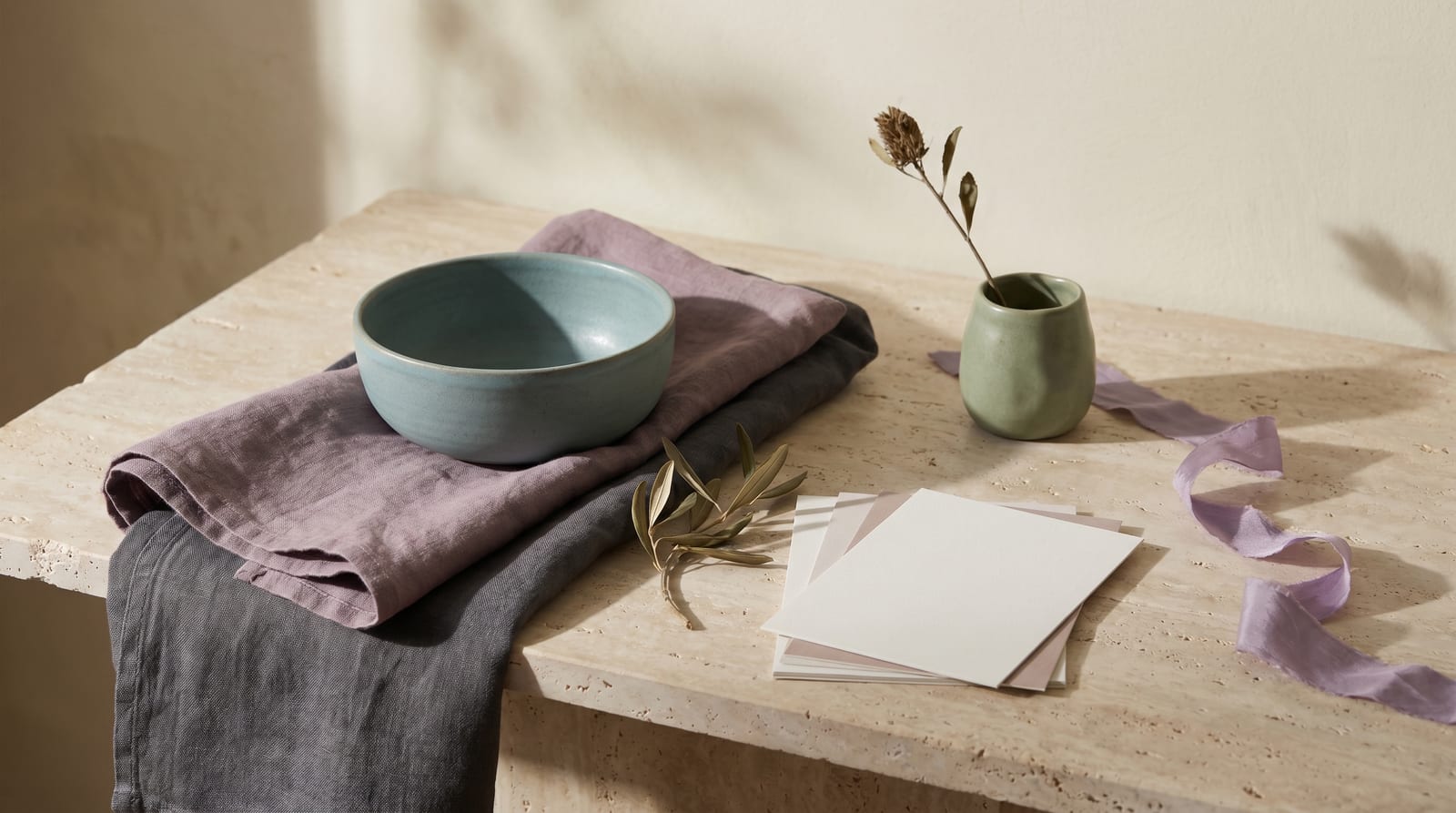 A soft summer editorial still-life: a tactile ensemble of folded linens, a small ceramic vessel, a stem of dried grass, and a pale stone on raw plaster, every element rendered in the palette's lavender-greys, dirty whites, and dusty roses.