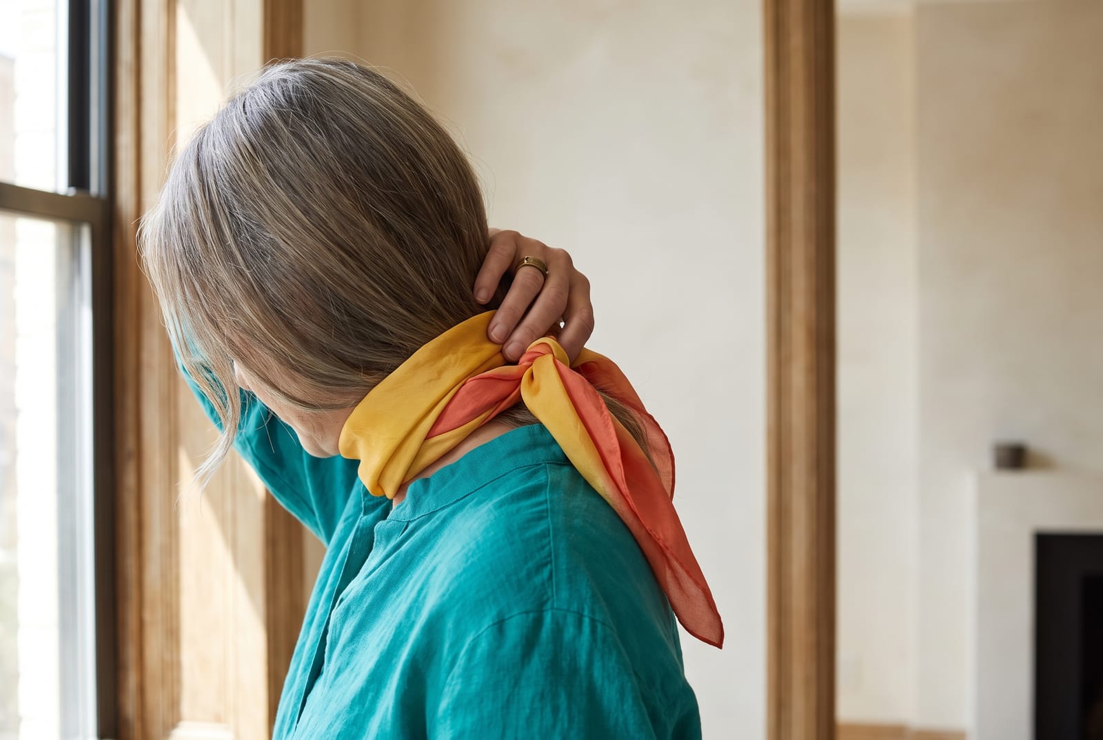 A warm spring portrait detail: a face-free editorial crop showing golden-blonde hair against a coral garment in bright, clear side-lit daylight, the kind of private, considered frame fashion editorials use when the face is out of view.